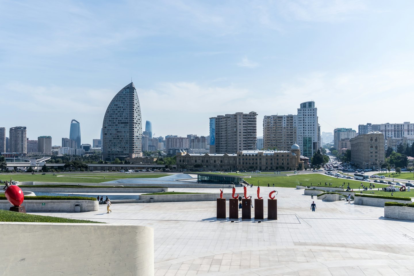 People walk through a city park with large red sculptures.
